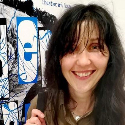 Nie wiem, kto jest na zdjęciu. Osoba z długimi, ciemnymi włosami uśmiecha się w pobliżu artystycznego plakatu.