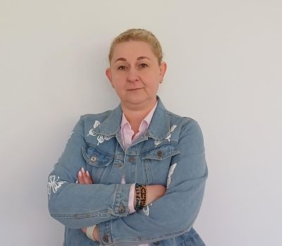 Kobieta z blond włosami stoi na białym tle, ubrana w dżinsową kurtkę i koszulę, z założonymi rękami.