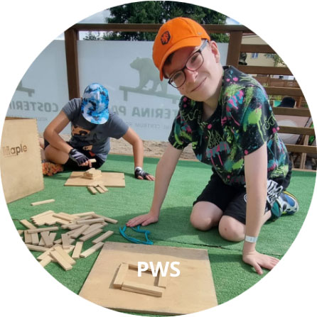 Dwoje dzieci bawi się drewnianymi klockami na zielonym dywanie na dworze. Jedno z nich uśmiecha się do kamery.